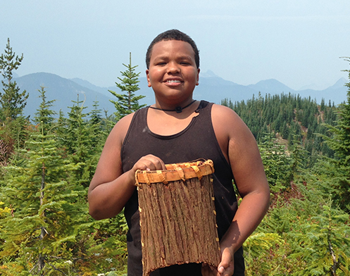 Sunny Kennebrew posing with his cedar basket standing in growing clearcut with the mountains in the background